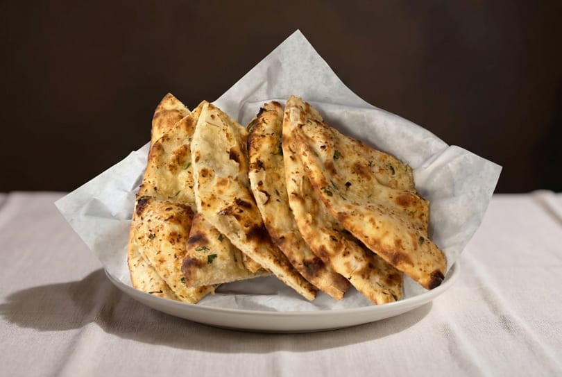 A white bowl filled with warm, golden-brown naan bread pieces, lined with white parchment paper.